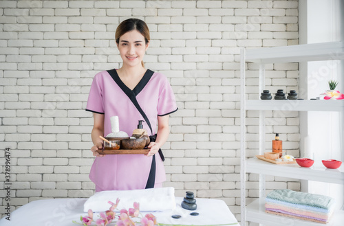 Happy young asian woman in uniform standing and holding tray of spa treatment accessories in spa massage room. Massage therapist female smile and welcome with service wellness for high quality.