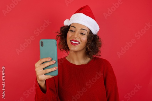 Isolated shot of pleased cheerful young woman with dark hair  makes selfie with mobile phone  dressed in casual sweater  isolated over beige background. People  technology and leisure concept