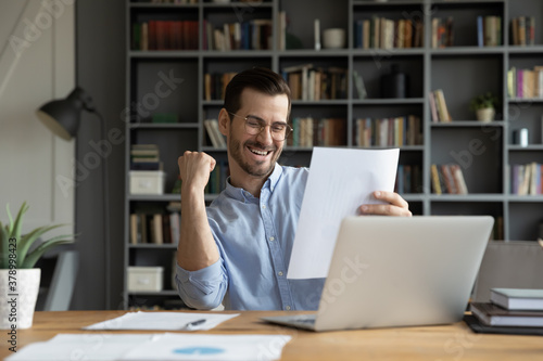 Excited young Caucasian man work on computer feel euphoric reading paper letter correspondence. Happy overjoyed male worker triumph with good pleasant news message in postal paperwork notice.