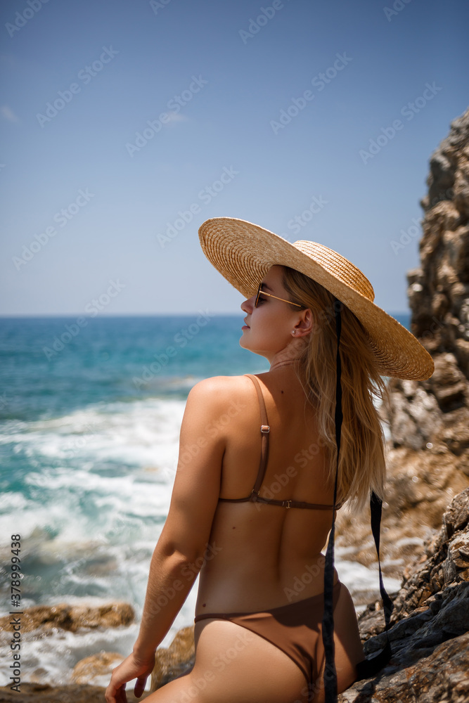 A beautiful girl in a swimsuit with a golden tan in a hat stands near the rocky coast. Summer sunny day by the sea. Vacation concept