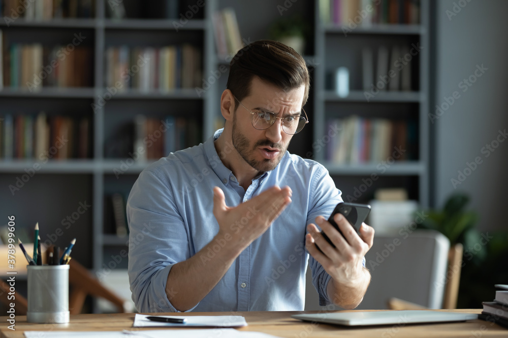 Mad millennial Caucasian male sit at desk look at cellphone shocked frustrated by gadget operational problems. Angry man stressed by message or error on smartphone, confused by slow internet. Mad millennial Caucasian male sit at desk look at cellphone shocked frustrated by gadget operational problems. Angry man stressed by message or error on smartphone, confused by slow internet.