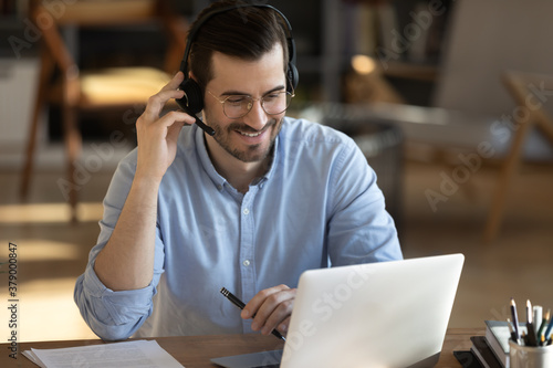 Smiling millennial Caucasian man in headset look at laptop screen work online from home office. Happy young man in headphones watch webinar take course or training on web. Distant education concept.