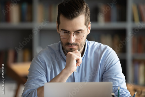Focused pensive young Caucasian man in glasses look at laptop screen work at home office online. Thoughtful serious millennial male freelancer browse surf internet on computer. Technology concept.