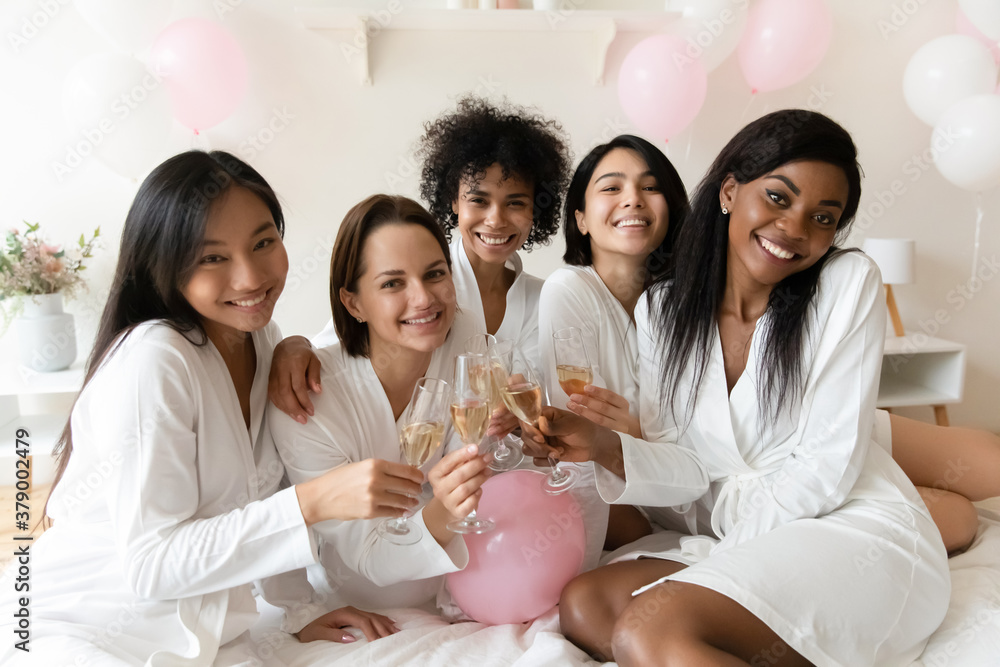 Portrait of smiling young mixed race pretty female best friends in white silk gowns, enjoying bachelorette hen pre-wedding party with sparkling wine, toasting saying wishes at luxury spa celebration. Portrait of smiling young mixed race pretty female best friends in white silk gowns, enjoying bachelorette hen pre-wedding party with sparkling wine, toasting saying wishes at luxury spa celebration.