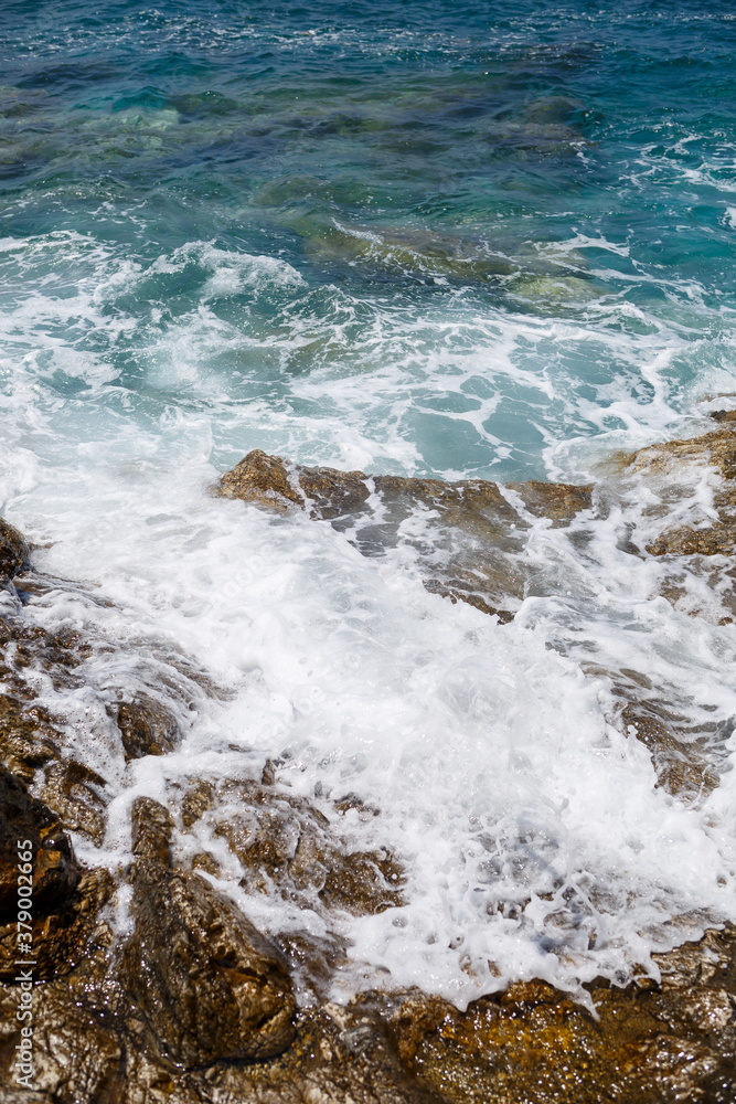 Wonderful views of the blue Mediterranean Sea. Sunny rocks, waves with foam and splashing water. The wave crashes into the rocks on the shore