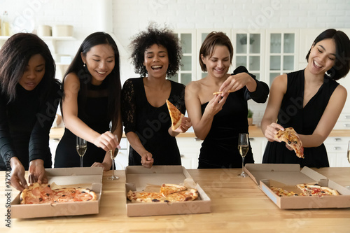 Happy young festive mixed race pretty women best friends in classic black dresses having fun at party, eating delivery italian fast food pizza, drinking champagne, enjoying meeting together indoors.