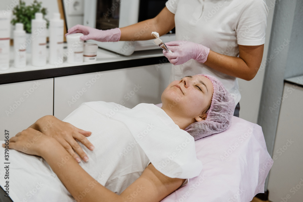 Professional Esthetician Apply Face Mask to the client's face in spa beauty center. Young woman getting facial care by beautician at spa salon Acne Treatment, face peeling mask Professional Esthetician Apply Face Mask to the client's face in spa beauty center. Young woman getting facial care by beautician at spa salon Acne Treatment, face peeling mask