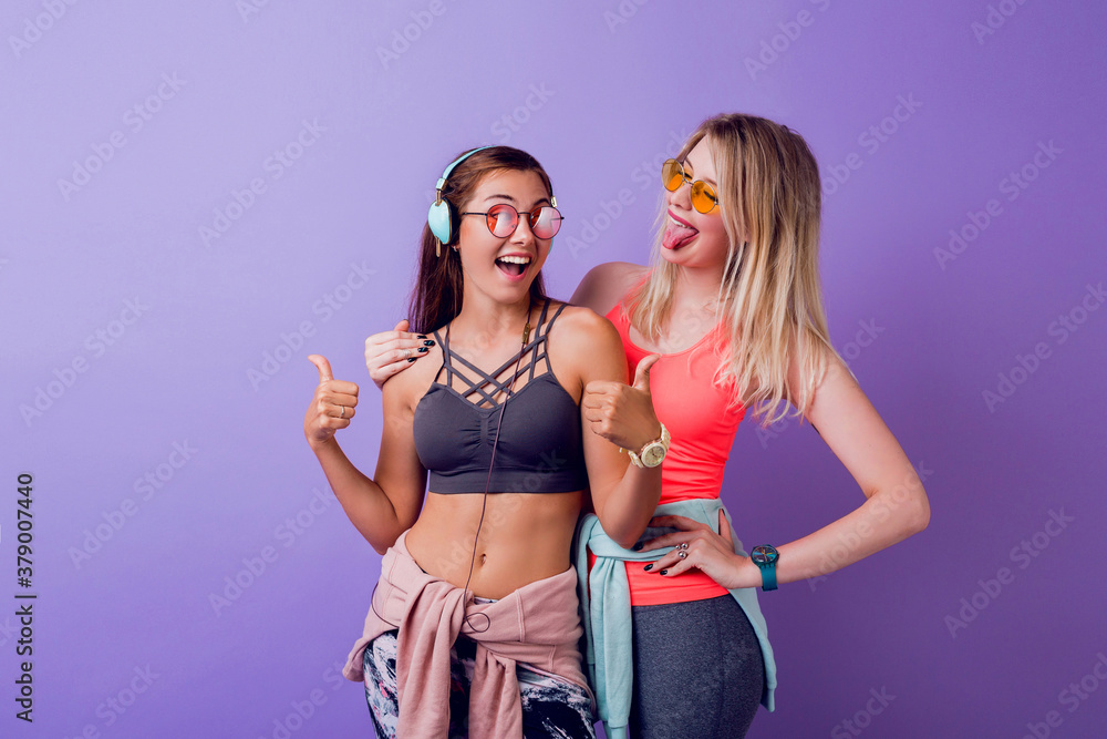 Two pretty girls in fashionable activewear posing on purple background in studio. Brunette and blond women standing together. Fitness and active fashion concept. Two pretty girls in fashionable activewear posing on purple background in studio. Brunette and blond women standing together. Fitness and active fashion concept.