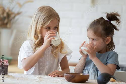 Two little sisters drinking eco milk from glasses at a messy, flour-covered table in kitchen. Happy 5 and 8-year-old siblings having a break and enjoying wholesome cow milk after making tasty cookies photo