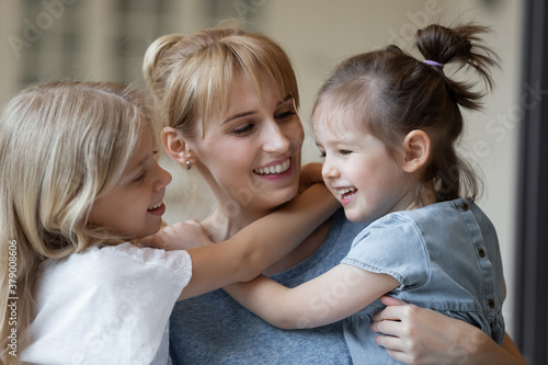 Close up of happy young mother in casual wear cuddling her two little daughters and smiling joyfully. Cute children embracing their mommy to express love and affection