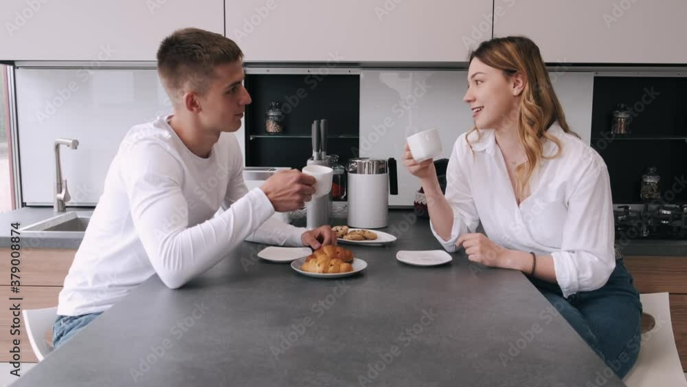 Beautiful young couple is talking. Pair looking at each other and smiling while eating and drinking in kitchen at home Beautiful young couple is talking. Pair looking at each other and smiling while eating and drinking in kitchen at home