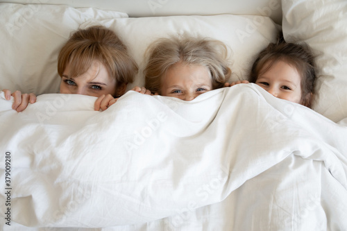 Billede på lærred Happy mom and children lying on comfortable bed together and peeking out from under soft white comforter