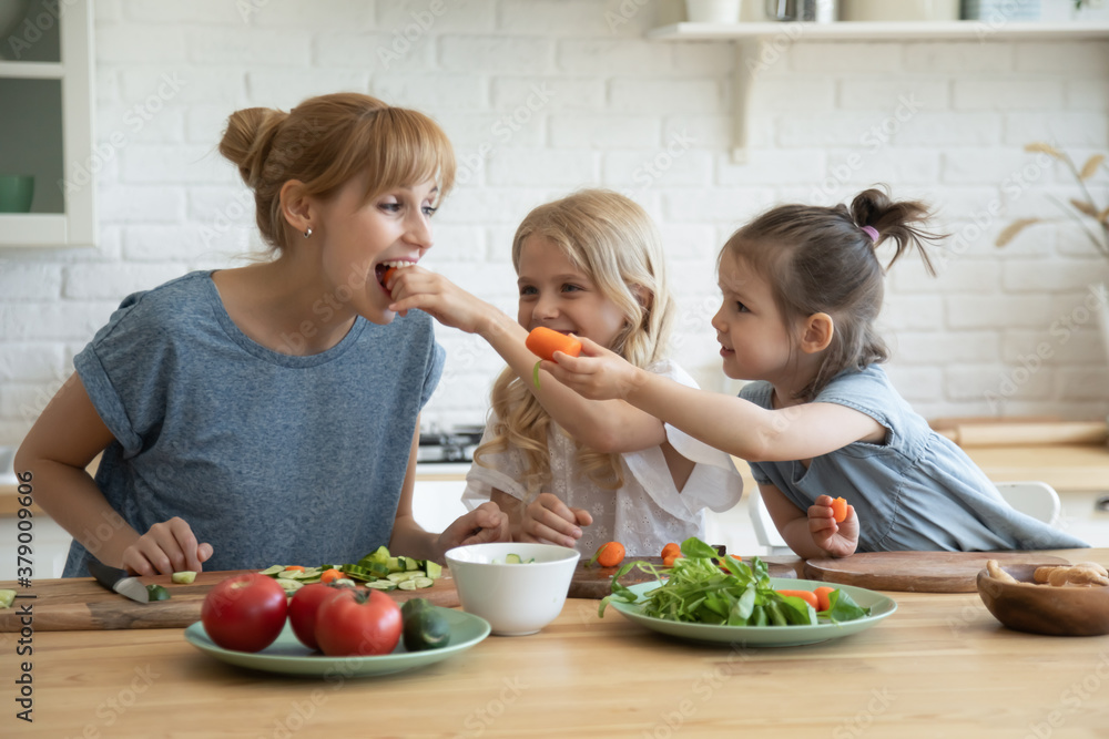 Playful kids feeding happy mum with carrots while chopping vegetables on cutting boards in the kitchen. Positive young mother teaching her 5 and 8 year old daughters to maintain healthy eating habits
