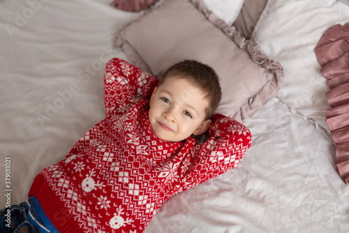 Happy little boy in red knitted sweater, dream on about presents laying in the bed. Christmas tree background. Enjoy winter holidays.