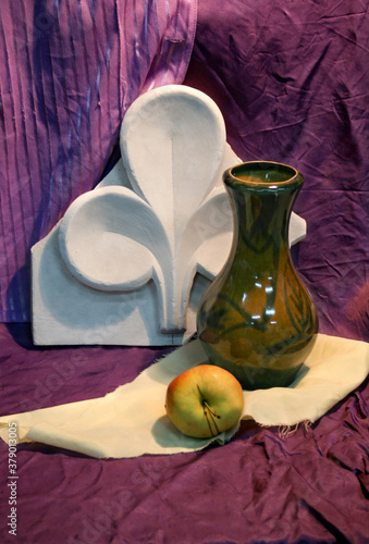 Artistic still life on a purple background with plaster floral ornament, apple and a bottle of olive color