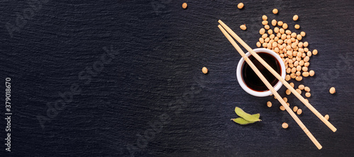 Soy sauce in a bowl with bamboo chopsticks and soya beans scattered on the black surface of the slate stone, banner, top view, closeup with space for text photo