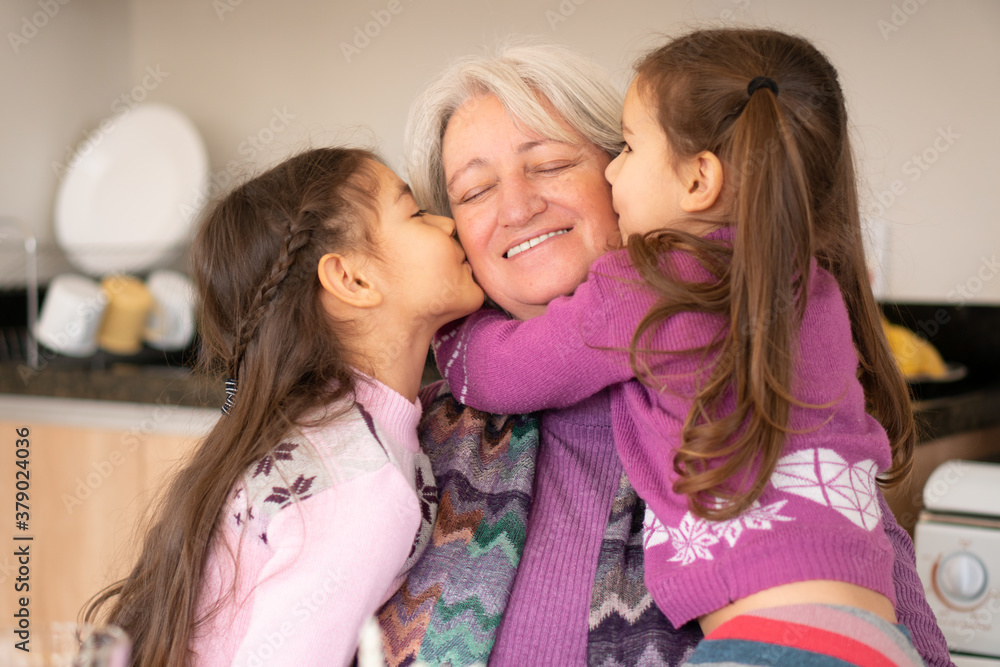 Cheerful brazilian young children and grandmother at tablekissing in the cheek. at kitchen table, inside. Unity, happiness, affection, love, care concept.