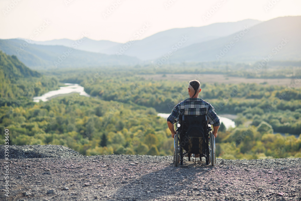Lonely young man in a wheelchair enjoying fresh air in sunny day on the mountain. People with disabilities travelling.
