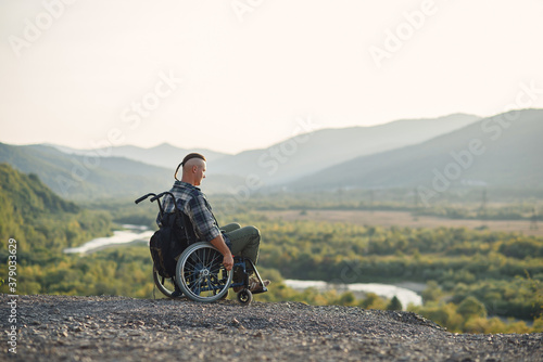 Lonely young man in a wheelchair enjoying fresh air in sunny day on the mountain. People with disabilities travelling.