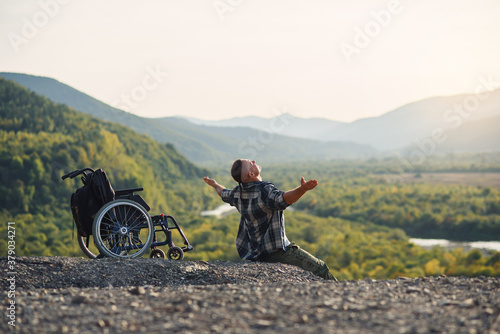Young disabled man sitting on the mountain near wheelchair and raised hands up enjoying fresh air and nature beauty. © gorynvd
