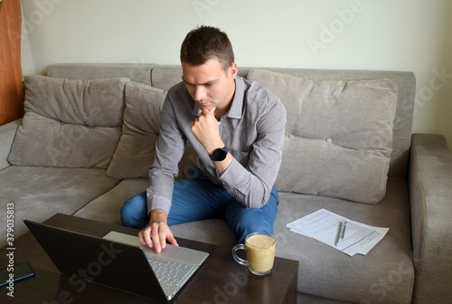 Young tired business man in shirt and trousers sitting on the couch and working with pensive serious face behind laptop after work in office. concept of a busy worker working at home. © Tatiana