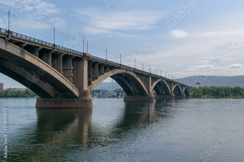 Old concrete bridge over a large river. Green trees on the shore, mountains. The background is the sky. Communal bridge in the city of Krasnoyarsk across the Yenisei River. © Sergei Tim