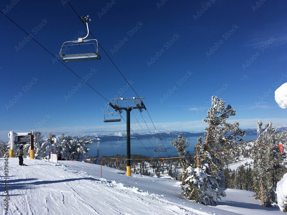 Winter landscape view of chair lift and Lake Tahoein the distance, as seen from the slopes of a ski resort, on a bluebird day with vibrant and clear blue skies Winter landscape view of chair lift and Lake Tahoein the distance, as seen from the slopes of a ski resort, on a bluebird day with vibrant and clear blue skies