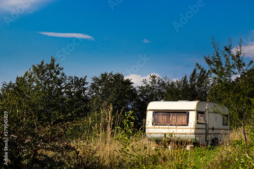Old white camper trailer parked on a grass field under blue sky with trees behind © medjutim