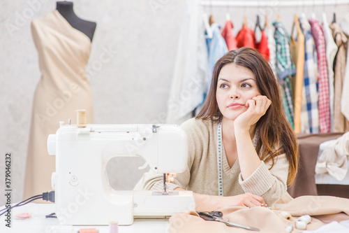 Thoughtful seamstress sits near a sewing machine and looks away