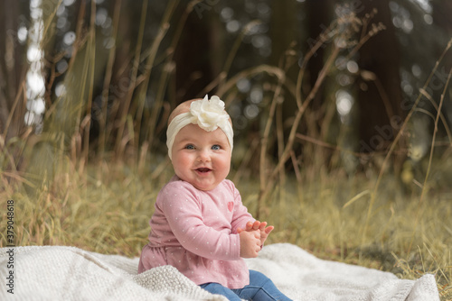 Adorable baby in pink shirt with white head band sitting smiling on blanket in park