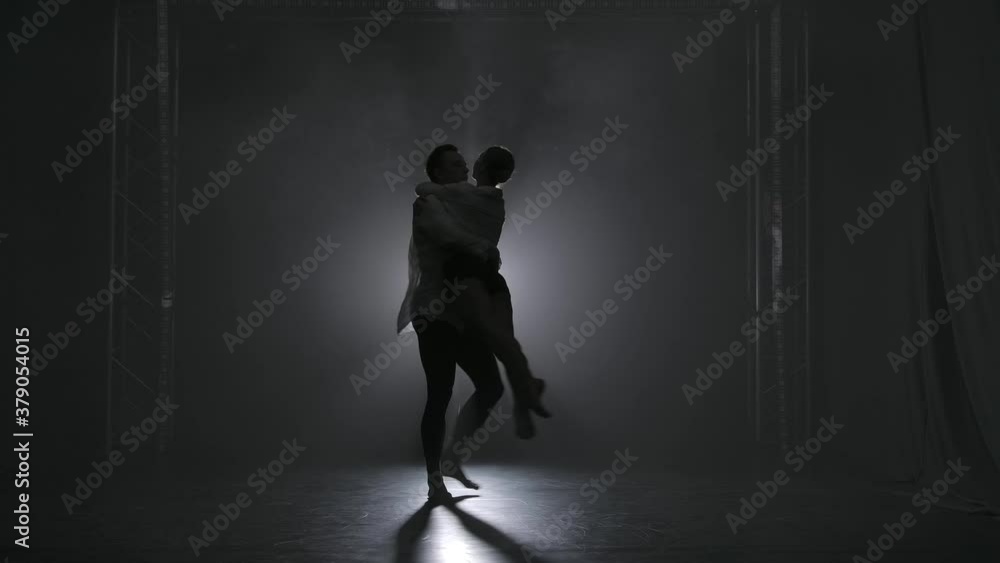 Silhouettes of a lovely couple of ballroom dancers performing in the spotlight. Sensual contemporary ballet choreography in a dark studio. Slow motion. Silhouettes of a lovely couple of ballroom dancers performing in the spotlight. Sensual contemporary ballet choreography in a dark studio. Slow motion.