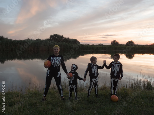 friendly family in carnival costumes of skeletons hold hands on river bank against background of sunset. Halloween Eve. Happy mom and three boys with pumpkins