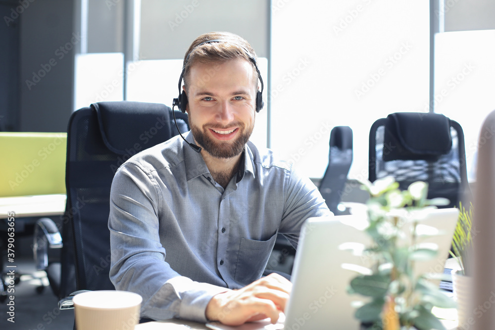 Smiling male call-center operator with headphones sitting at modern office, consulting online information in a laptop, looking up information in a file in order to be of assistance to the client.
