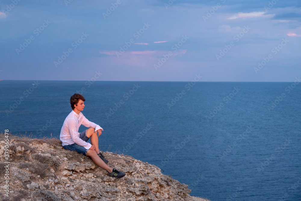 A young man sits on a rocky seashore at sunset.