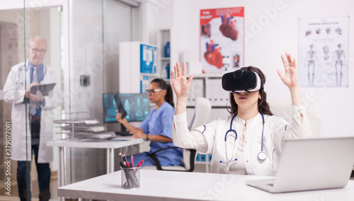Young medic doing hand gesture wearing virtual reality headset in hospital office. Doctor in white coat with stethoscop. Senior physician writing notes on clipboard. Nurse in blue uniform looking at x