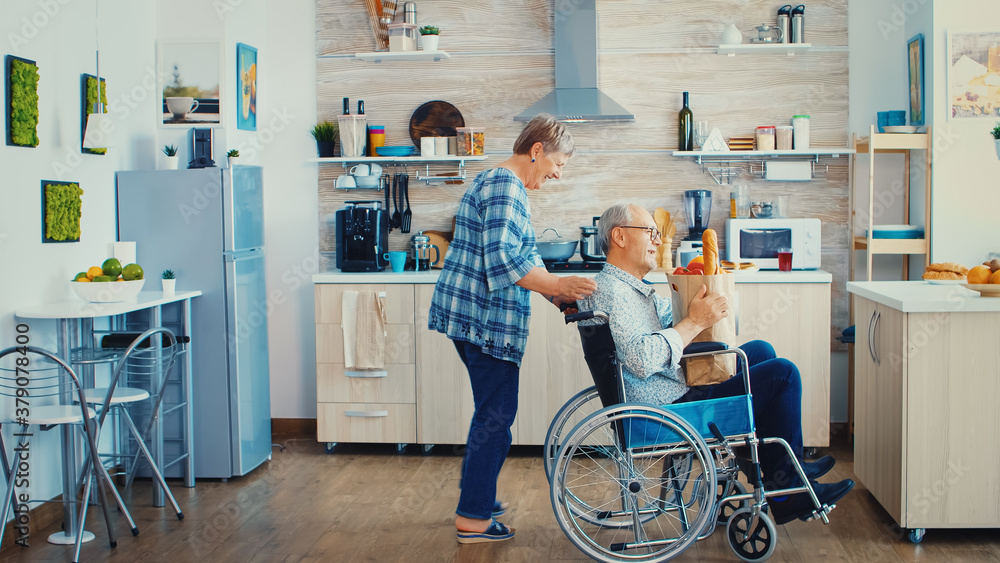 Fototapeta premium Old woman pushing invalid senior husband in wheelchair after arriving with grocery paper bag from supermarket. Mature people with fresh vegetables for cooking breakfast.
