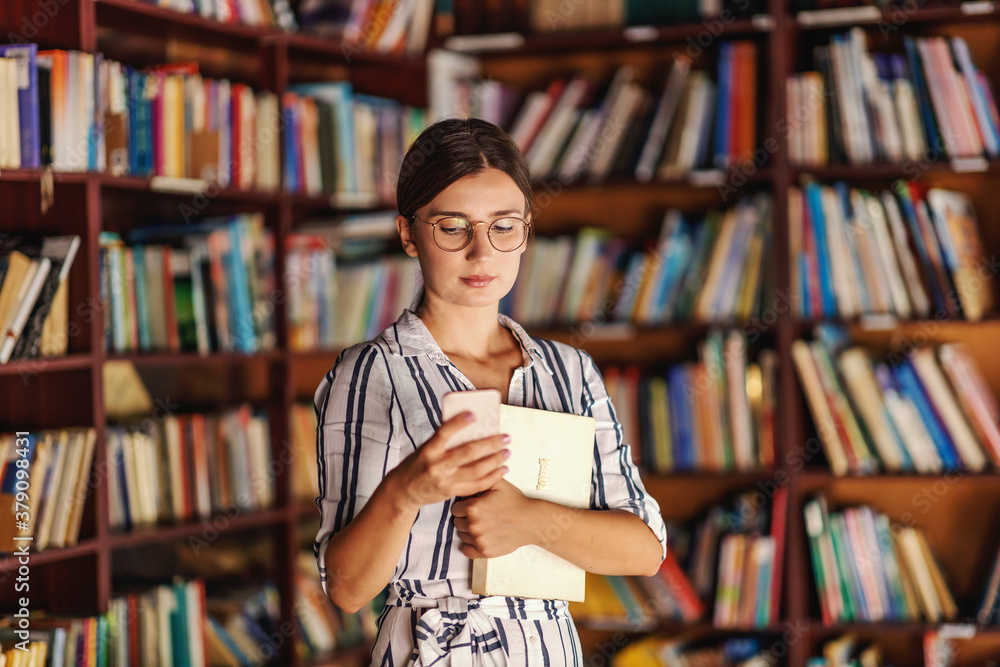 Young attractive college girl standing in library, holding a book and using smart phone. Young attractive college girl standing in library, holding a book and using smart phone.