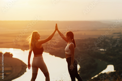 Happy girls doing sport at sunset in the mountains. They give each other high five standing on mountain under evening sky with sunset and landscape of river.
