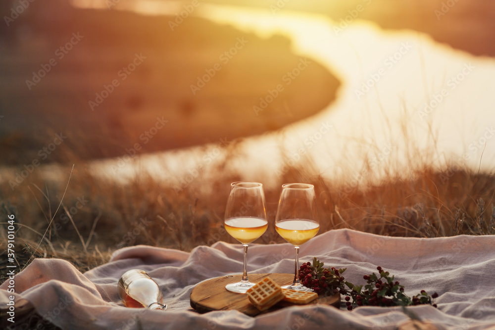 Romantic picnic on the mountain with river and sunset on background. Bottle of wine, glasses, plaid and dessert.