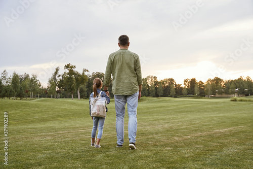 Rear view of father and little daughter standing in a green field on a warm day, walking together in the park