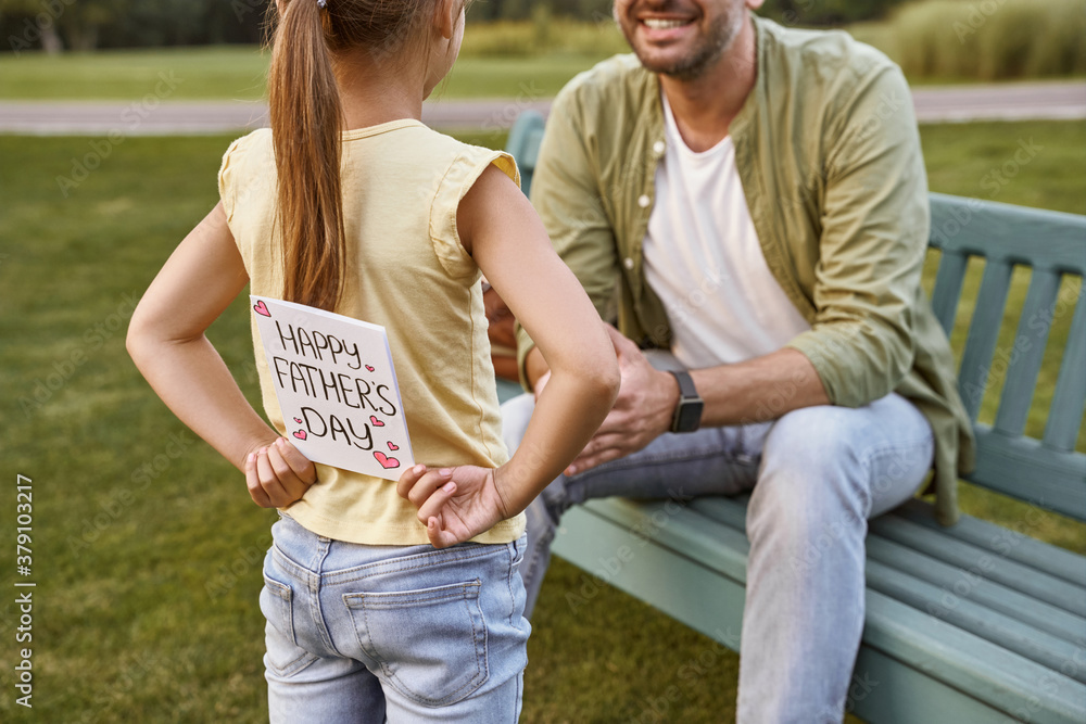 Naklejka premium Father sitting on the wooden bench in park while his little daughter hiding handmade postcard for him, small girl making a surprise for Happy Fathers Day