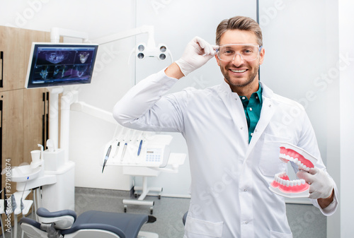Portrait of a handsome dentist man smiling and looking at the camera, holding a tooth model. Painless caries treatment in a modern dental office
