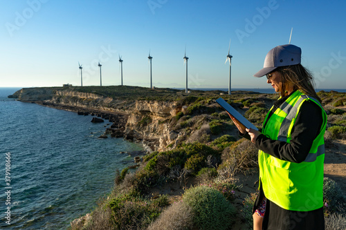 Female engineer standing with digital tablet against wind turbine on sunny day, looking at wind turbine
