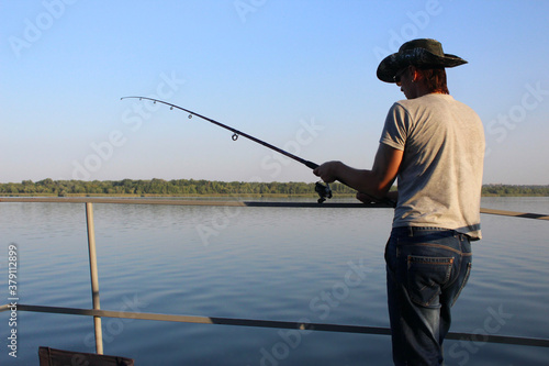  fisherman in a hat with a fishing rod in his hands stot on the river bank view from the back