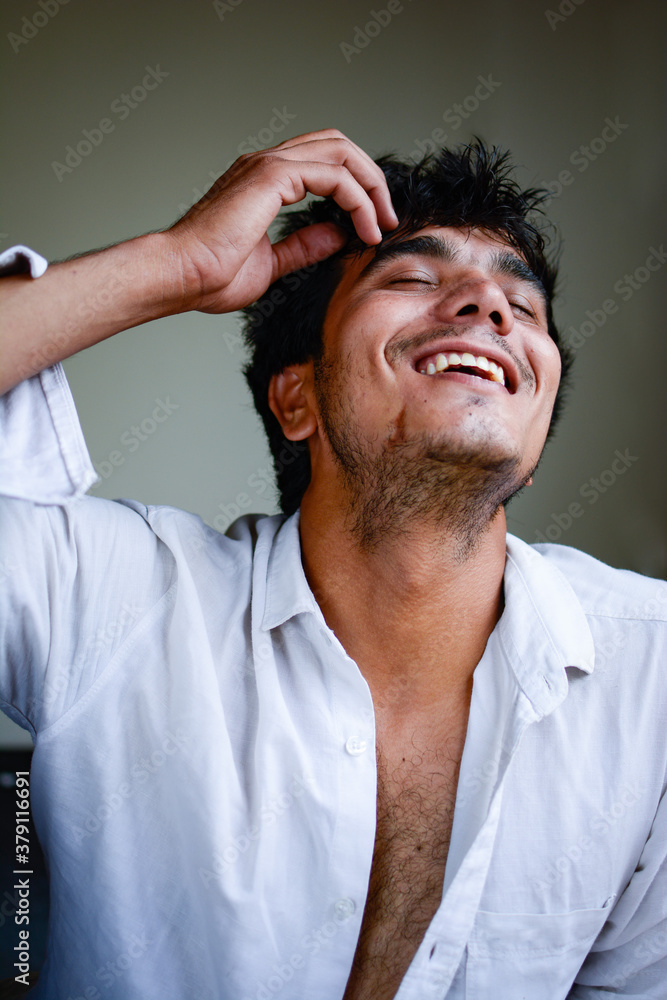 Obraz premium indoor portrait shot of a young handsome Indian man wearing a white unbuttoned shirt and laughing. he has a hairy chest. one hand is lifted