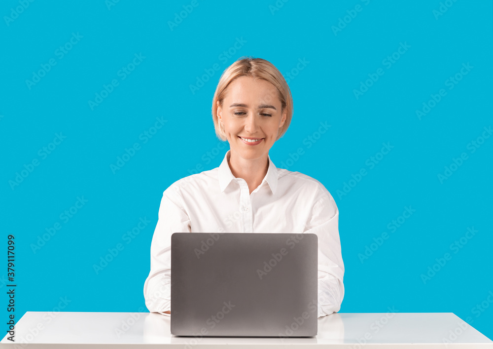 Positive business lady using laptop computer at her desk on blue studio background Positive business lady using laptop computer at her desk on blue studio background