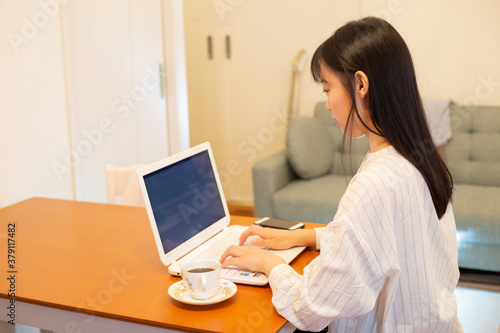 リモートワーク。自宅のキッチンテーブルでノートパソコンでの仕事中、コーヒーを飲む若く美しいビジネスウーマン。