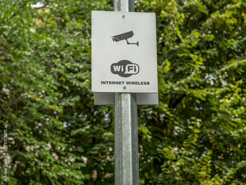 A sign on a metal pole in a park with Wi Fi internet wireless and a security camera