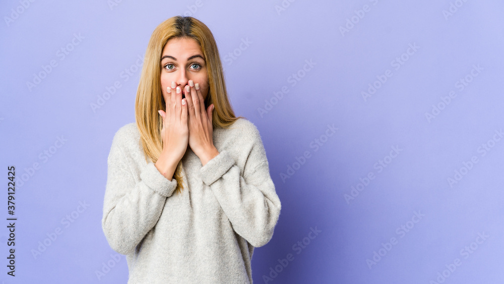 Young blonde woman isolated on purple background shocked, covering mouth with hands, anxious to discover something new. Young blonde woman isolated on purple background shocked, covering mouth with hands, anxious to discover something new.