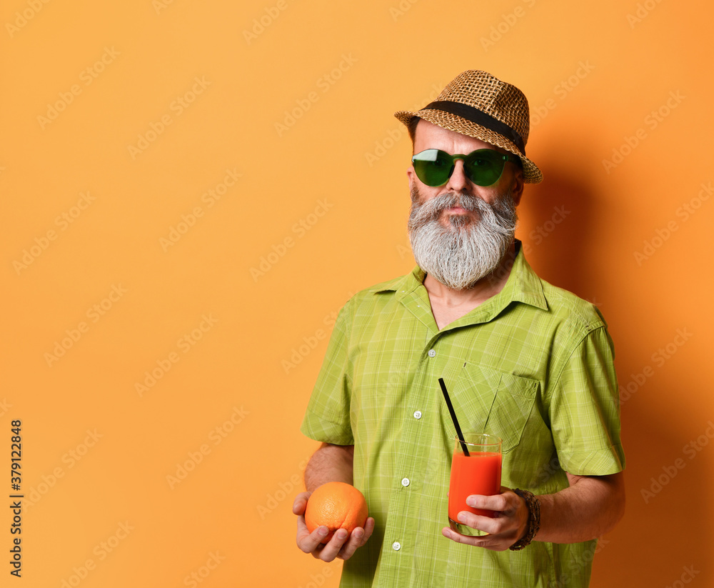 handsome man in a hat holds an orange and a glass of freshly squeezed juice with a black tube, posing sideways on an orange studio background. Close copy space handsome man in a hat holds an orange and a glass of freshly squeezed juice with a black tube, posing sideways on an orange studio background. Close copy space